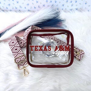 May include: Clear rectangular bag with a maroon trim and a patterned strap. The bag has the text "TEXAS A&M Aggies" in maroon and white. The strap is maroon and white with a geometric design. A gold-colored clasp is visible.