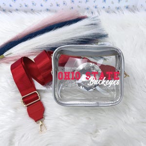 May include: A clear, square crossbody bag with a red strap and gold-colored hardware. The bag features the text "OHIO STATE Buckeyes" in red and white. A large, clear, diamond-shaped embellishment is inside the bag.