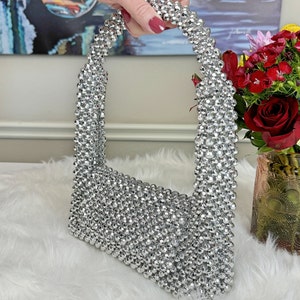 May include: A silver beaded handbag with a unique, rounded shape. The bag is made entirely of small, silver beads that are tightly woven together.