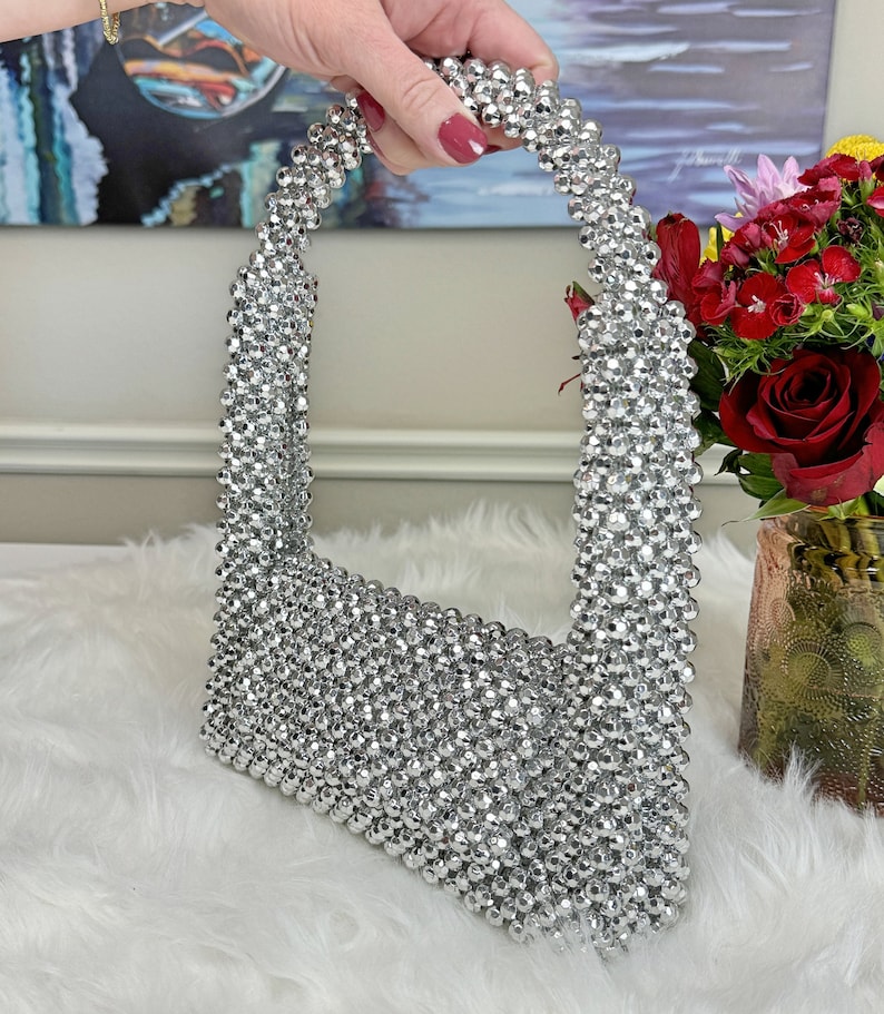 May include: A silver beaded handbag with a unique, rounded shape. The bag is made entirely of small, silver beads that are tightly woven together.