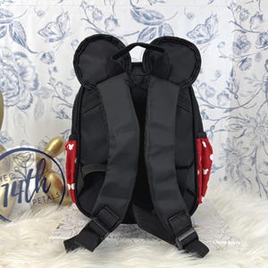 May include: A black backpack featuring two rounded ears at the top, adjustable shoulder straps, and red side pockets with a white polka dot pattern. The design includes a top handle and black nylon straps with plastic buckles for secure closure.