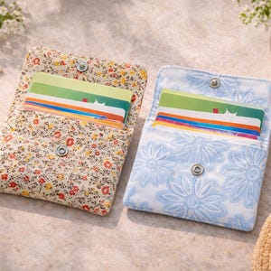 Peut inclure: Deux porte-cartes rectangulaires en tissu avec fermetures à pression en métal. Le porte-cartes de gauche présente un petit motif floral multicolore sur fond crème. Celui de droite affiche un grand motif floral bleu clair et blanc.