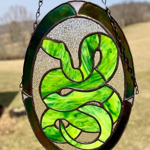 Puede incluir: Una ventana de vitral colgada de una cadena, con un diseño de serpiente verde con un borde marrón.