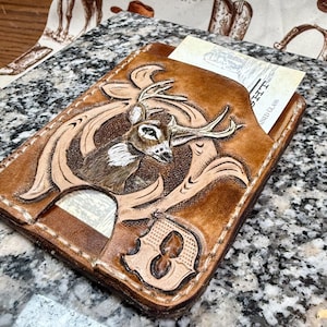 May include: Hand-tooled leather wallet with a deer design. The brown wallet features a detailed carving of a deer head with antlers, surrounded by an ornate border. A card is visible in the front pocket. The wallet is stitched with white thread.