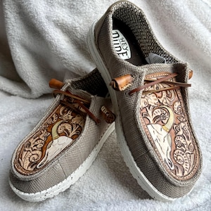 Könnte beinhalten: Ein Paar Slip-On-Schuhe mit Obermaterial aus beigefarbenem Canvas und weiß gesprenkelten Sohlen. Die Schuhspitze ziert ein detailliertes, geschnitztes Lederdesign eines Widder-Schädels und floralen Elementen. Braune Lederschnürsenkel und Akzente.