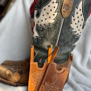 Könnte beinhalten: Zwei Handyhüllen aus Leder in Braun- und Orangetönen mit weißen Stickmustern. Sie werden vor einem abgenutzten Cowboy-Stiefel mit einem Flügeldesign und Sternaussparungen präsentiert.