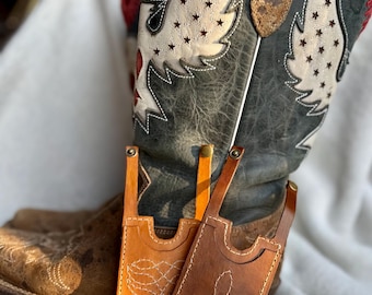 Op de afbeelding: Twee leren telefoonhoesjes in bruin- en oranjetinten, met wit gestikte ontwerpen. Ze worden getoond voor een versleten cowboylaars met een vleugelontwerp en steruitsparingen.
