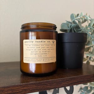 Puede incluir: Una vela en tarro de cristal marrón con tapa negra y una etiqueta que dice "purify candle co." y un versículo bíblico. La vela está sobre una estantería de madera marrón oscuro junto a una pequeña planta en maceta negra.