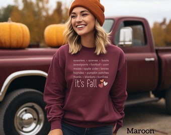 May include: A maroon sweatshirt with the text "it's fall" and a list of fall activities, including sweaters, scarves, boots, sweatpants, football, corn, mazes, apple cider, leaves, hayrides, pumpkin patches, thanks, blankets, and bonfires. Three small hearts are next to the text "it's fall".