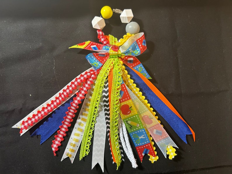 May include: A vibrant handmade ribbon lei with ribbons in red, blue, orange, and green. The lei features a bow and decorative elements, including a yellow bead and wooden shapes. The ribbons have school-themed patterns.