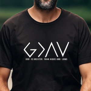 May include: A man wearing a black t-shirt with a white graphic that says "G>A>V" and "GOD IS GREATER THAN HIGHS AND LOWS". He is wearing a brown hat and has a beard.