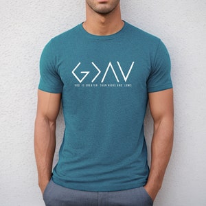 May include: A teal blue t-shirt with a white graphic design of the letters "G>A<V" and the text "God is greater than highs and lows".
