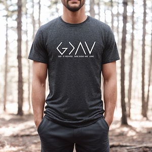 May include: A dark gray t-shirt with a white graphic design that reads "G>A<V" and "God is greater than highs and lows".