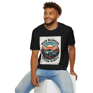 May include: A black t-shirt featuring a vintage-style graphic of a jeep driving through a mountainous landscape. The graphic reads "Mud Slinger Gettin' Dirty".