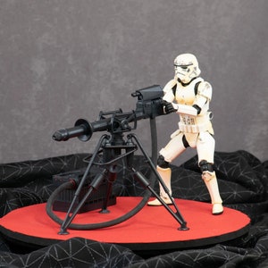 Puede incluir: Una figura de acción de Stormtrooper blanca sosteniendo un cañón blaster negro en un trípode. El cañón está conectado a una caja negra con una manguera. La figura y el cañón están en una plataforma circular roja.