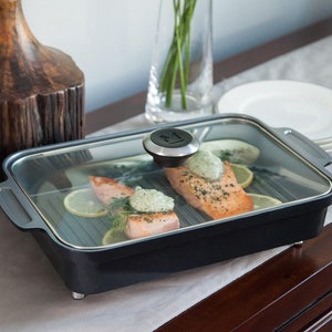 May include: A black rectangular grill pan with a glass lid, containing cooked salmon fillets, lemon slices, and herbs. The pan has a silver handle and a black knob. The food is arranged on a gray surface, with a vase and plates in the background.