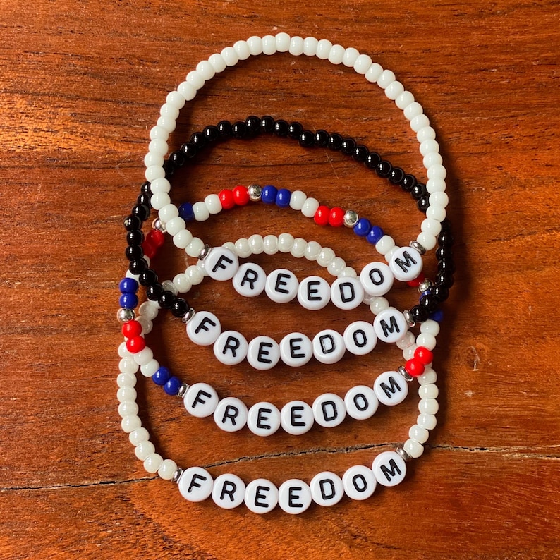 May include: Four beaded bracelets with the word "FREEDOM" spelled out in white letter beads. The bracelets are white, black, red, and blue. The bracelets are on a wooden surface.