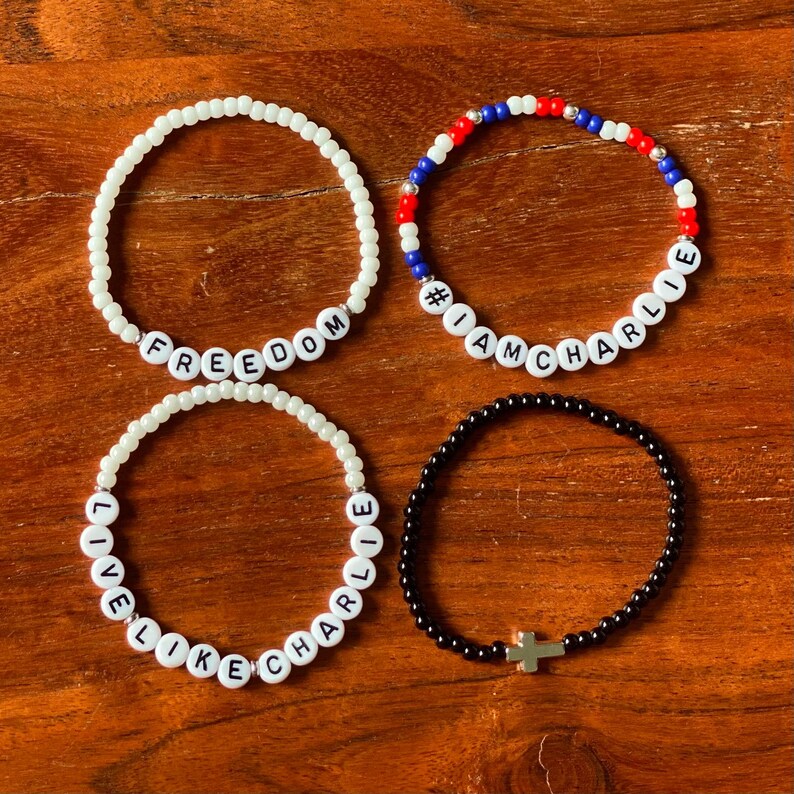 May include: Four beaded bracelets on a wooden surface. One white bracelet spells "FREEDOM", another with red, white, and blue beads says "#IAMCHARLIE", and a third says "LIVE LIKE CHARLIE". The fourth bracelet is black with a gold cross.