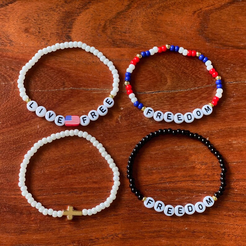 May include: Four beaded bracelets in the image. One white bracelet has the words "LIVE FREE" and an American flag bead. Another bracelet has red, white, and blue beads with the word "FREEDOM". The third bracelet is white with a gold cross. The last bracelet is black with the word "FREEDOM".