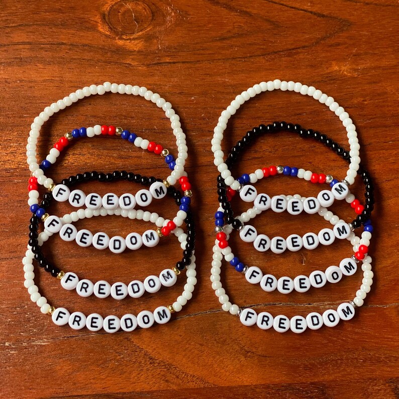 May include: Several beaded bracelets with the word "FREEDOM" spelled out in white letter beads. The bracelets are in various colors, including white, black, red, blue, and gold. The bracelets are displayed on a wooden surface.