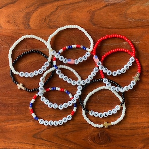 May include: A collection of beaded bracelets in red, white, black, and blue. The bracelets feature white letter beads spelling out phrases like "FREEDOM", "WORK HARDER", and "LIVE FREE". Some bracelets include cross charms and American flag beads.