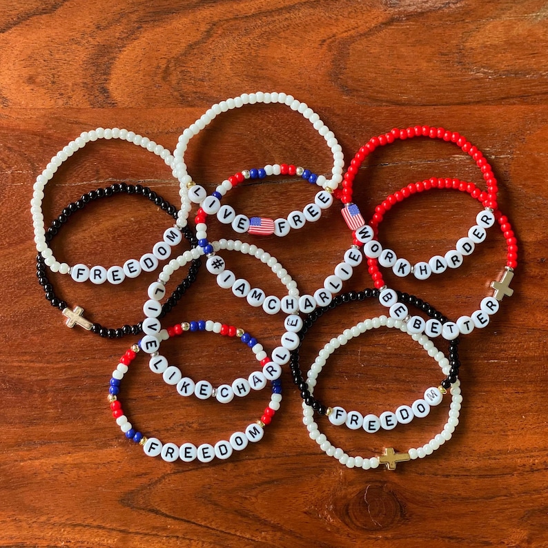 May include: A collection of beaded bracelets in red, white, black, and blue. The bracelets feature white letter beads spelling out phrases like "FREEDOM", "WORK HARDER", and "LIVE FREE". Some bracelets include cross charms and American flag beads.