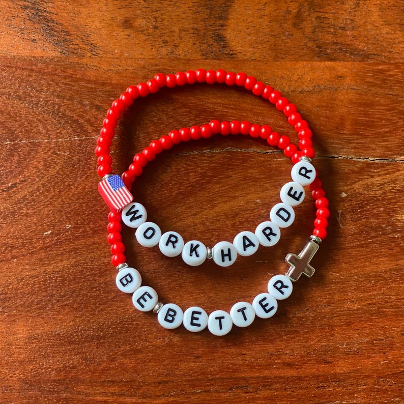 May include: Two red beaded bracelets. One bracelet has the text "WORK HARDER" spelled out in white letter beads. The other bracelet has the text "BE BETTER" spelled out in white letter beads. One bracelet has a small American flag charm and the other has a cross charm.