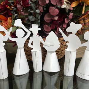 May include: Seven white paper figures of people, each with a different pose and holding a different object, stand on white cone-shaped bases. The figures are arranged in a row on a table with a blurred background of a wicker basket and autumn foliage.