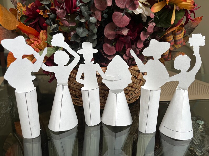 May include: Seven white paper figures of people, each with a different pose and holding a different object, stand on white cone-shaped bases. The figures are arranged in a row on a table with a blurred background of a wicker basket and autumn foliage.