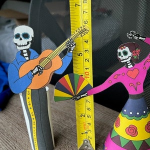 May include: Two paper cutouts of skeletons dressed in traditional Mexican Day of the Dead costumes. The skeleton on the left is playing a guitar and the skeleton on the right is holding a colorful fan. The skeletons are standing on a wooden surface with a measuring tape behind them.