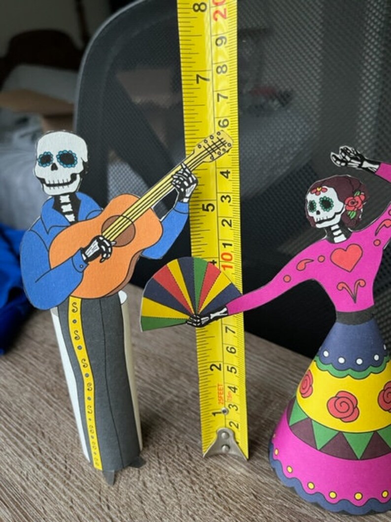 May include: Two paper cutouts of skeletons dressed in traditional Mexican Day of the Dead costumes. The skeleton on the left is playing a guitar and the skeleton on the right is holding a colorful fan. The skeletons are standing on a wooden surface with a measuring tape behind them.