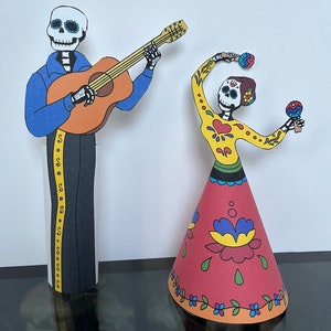 May include: Two paper cutouts of skeletons dressed in traditional Mexican Day of the Dead costumes. The skeleton on the left is playing a guitar and the skeleton on the right is holding maracas. Both skeletons have colorful floral patterns on their clothing.