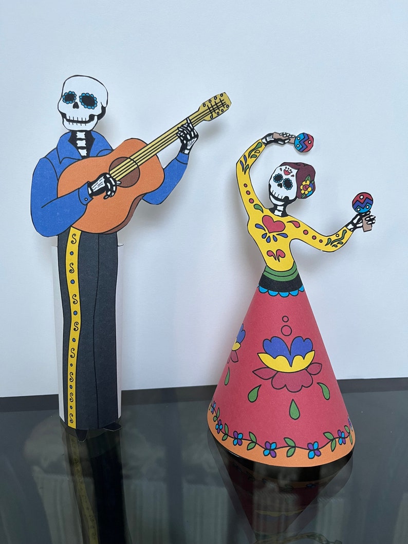 May include: Two paper cutouts of skeletons dressed in traditional Mexican Day of the Dead costumes. The skeleton on the left is playing a guitar and the skeleton on the right is holding maracas. Both skeletons have colorful floral patterns on their clothing.
