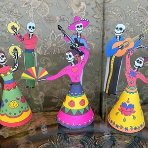 May include: Five paper cutouts of skeletons dressed in traditional Mexican Day of the Dead costumes. The skeletons are playing musical instruments, dancing, and holding a fan. The cutouts are colorful and feature intricate designs.