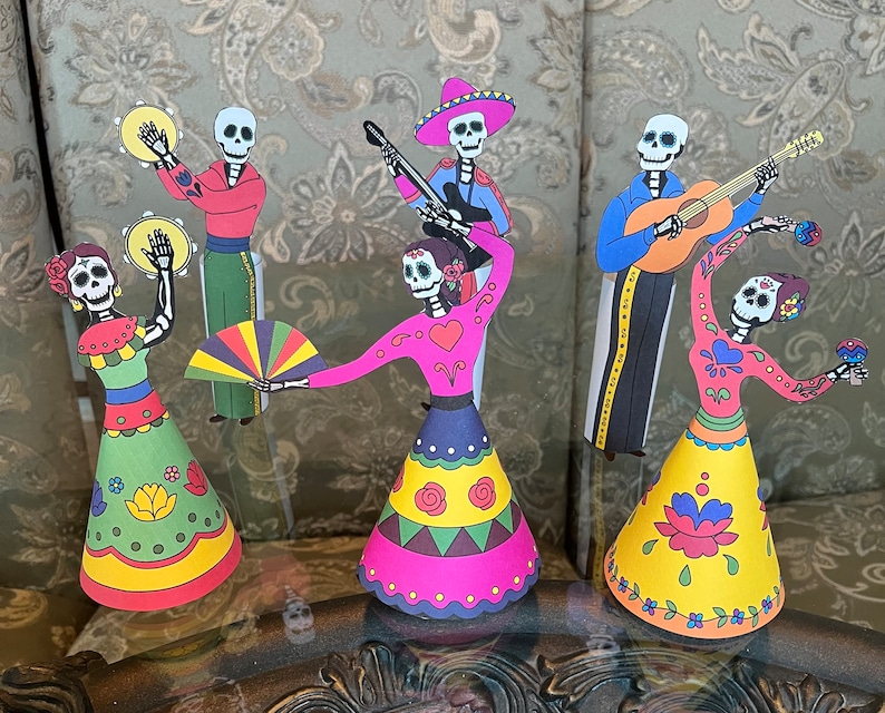 May include: Five paper cutouts of skeletons dressed in traditional Mexican Day of the Dead costumes. The skeletons are playing musical instruments, dancing, and holding a fan. The cutouts are colorful and feature intricate designs.