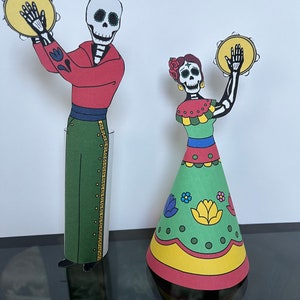 May include: Two paper cutouts of skeletons dressed in traditional Mexican Day of the Dead costumes. The male skeleton is wearing a red shirt, green pants, and a black sash. The female skeleton is wearing a green dress with red, yellow, and blue accents. Both skeletons are holding yellow tambourines.