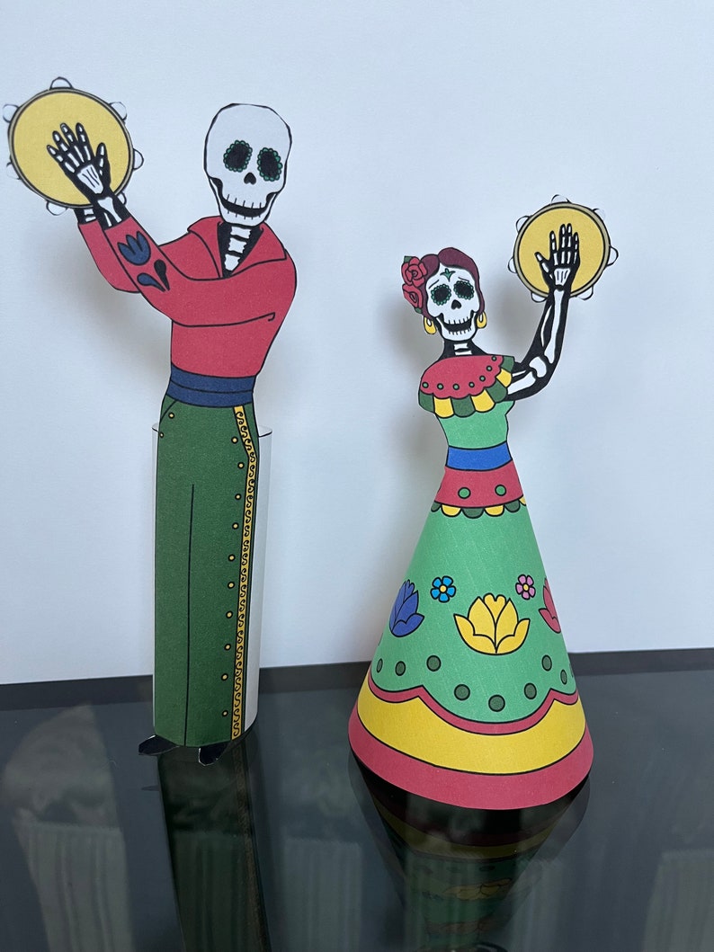 May include: Two paper cutouts of skeletons dressed in traditional Mexican Day of the Dead costumes. The male skeleton is wearing a red shirt, green pants, and a black sash. The female skeleton is wearing a green dress with red, yellow, and blue accents. Both skeletons are holding yellow tambourines.