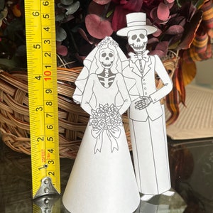 May include: A black and white paper craft of a skeleton bride and groom. The bride is wearing a long white dress and a veil. The groom is wearing a tuxedo and a top hat. The couple are standing on a table with a measuring tape in the background.