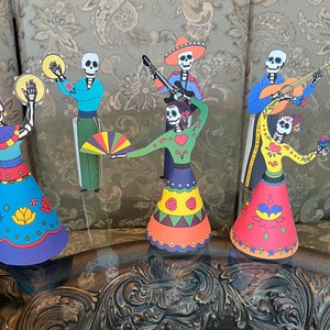 May include: A set of six Day of the Dead paper figures, each with a unique design and colorful clothing. The figures include a musician playing a guitar, a musician playing a tambourine, a dancer with a fan, and three dancers with flowers.