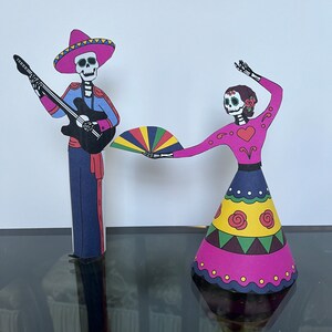 May include: Two paper cutouts of skeletons dressed in traditional Mexican Day of the Dead costumes. The male skeleton is playing a guitar and wearing a sombrero. The female skeleton is holding a colorful fan and wearing a pink dress with yellow and green accents.