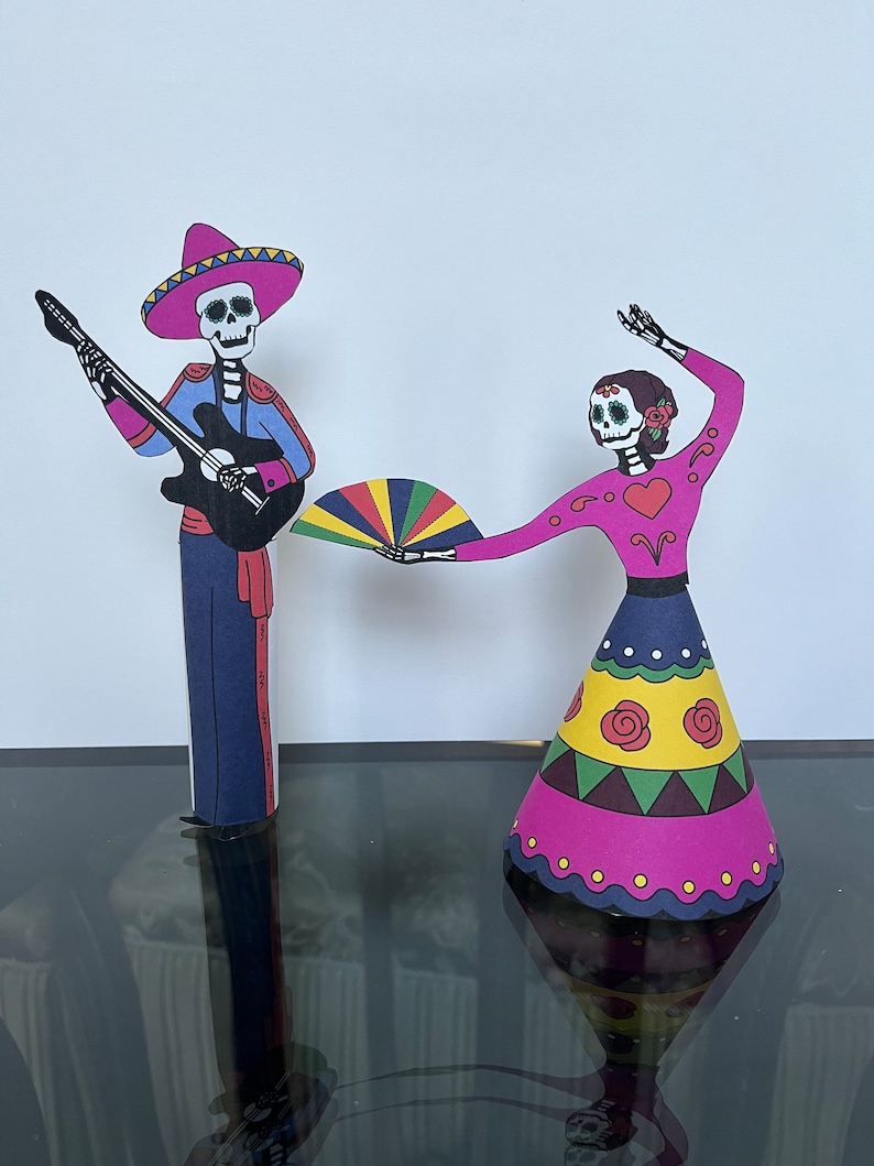 May include: Two paper cutouts of skeletons dressed in traditional Mexican Day of the Dead costumes. The male skeleton is playing a guitar and wearing a sombrero. The female skeleton is holding a colorful fan and wearing a pink dress with yellow and green accents.