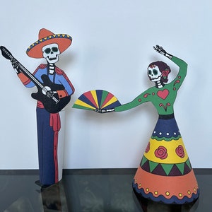 May include: Two paper cutouts of skeletons dressed in traditional Mexican Day of the Dead costumes. The male skeleton is playing a guitar and wearing a sombrero. The female skeleton is holding a colorful fan and wearing a green dress with a red heart.