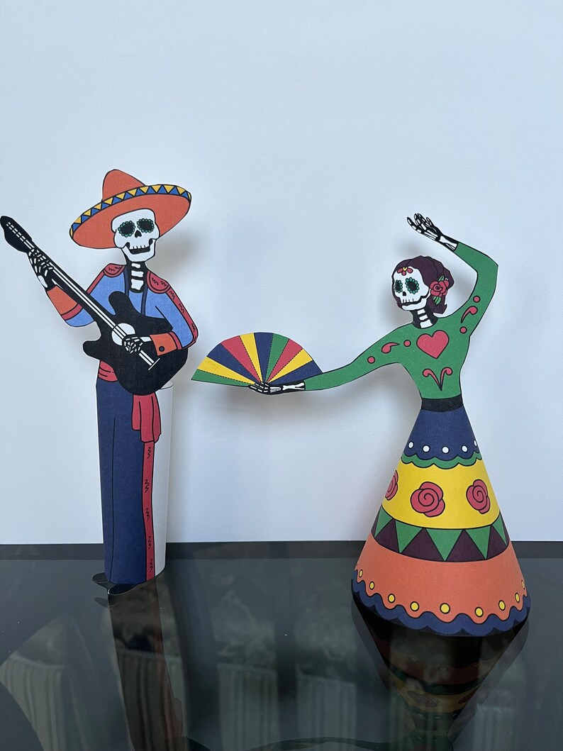 May include: Two paper cutouts of skeletons dressed in traditional Mexican Day of the Dead costumes. The male skeleton is playing a guitar and wearing a sombrero. The female skeleton is holding a colorful fan and wearing a green dress with a red heart.