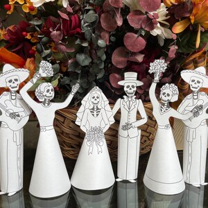May include: Five paper cut-outs of skeletons dressed in wedding attire. The skeletons are holding flowers and a guitar. The skeletons are standing in front of a bouquet of flowers.