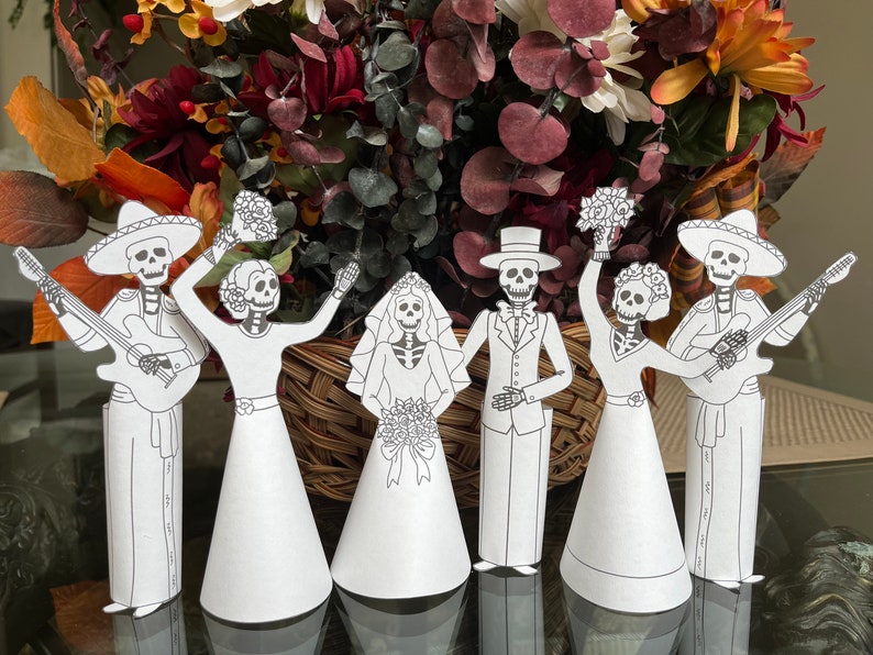 May include: Five paper cut-outs of skeletons dressed in wedding attire. The skeletons are holding flowers and a guitar. The skeletons are standing in front of a bouquet of flowers.