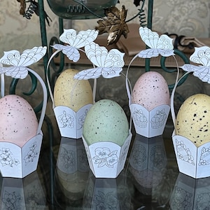 May include: Six white paper Easter baskets with butterfly cutouts and handles. Each basket holds a speckled Easter egg in different colours: pink, green, and yellow.