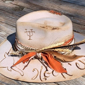 May include: A white cowboy hat with a brown leather band and a feather accent. The hat has a distressed look and the words "You're gonna love this" are written on the brim.