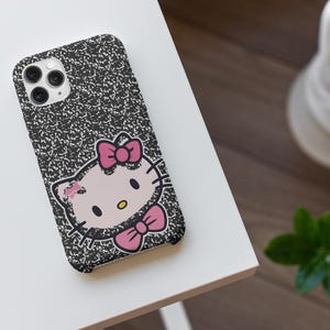 以下が含まれることがあります： Hello Kittyのグラフィックが特徴の、白黒のコンポジションノートスタイルの携帯電話ケース。ケースにはピンクのリボンとピンクの蝶ネクタイが付いています。携帯電話ケースは白い面にあります。
