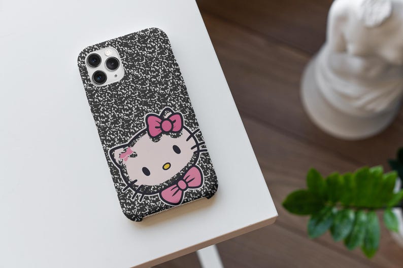以下が含まれることがあります： Hello Kittyのグラフィックが特徴の、白黒のコンポジションノートスタイルの携帯電話ケース。ケースにはピンクのリボンとピンクの蝶ネクタイが付いています。携帯電話ケースは白い面にあります。