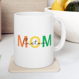 May include: White ceramic mug with the word "MOM" in orange, yellow, and green block letters. The word "Custom" is written in a cursive font between the letters. The mug sits on a beige coaster.